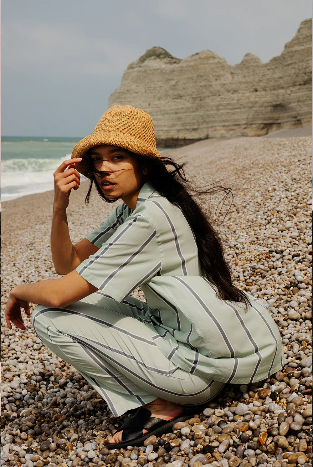 pantalon et chemise bleus à rayures portés par une femme accroupie sur une plage de galet en Normanddie